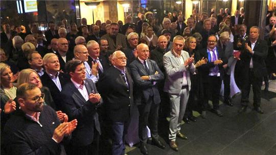 Große Gruppe von Menschen in formeller Kleidung, die in einem Innenraum stehen und applaudieren.