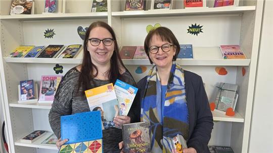 Zwei Frauen stehen vor einem Bücherregal mit verschiedenen Büchern und halten jeweils mehrere Bücher und ein Spielbrett beziehungsweise ein Spielzeug in den Händen.