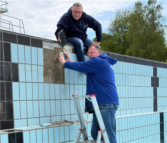 Zwei Männer reparieren eine hellblaue geflieste Wand, einer steht auf einer Leiter, der andere sitzt auf der Wand und bearbeitet die Fugen.