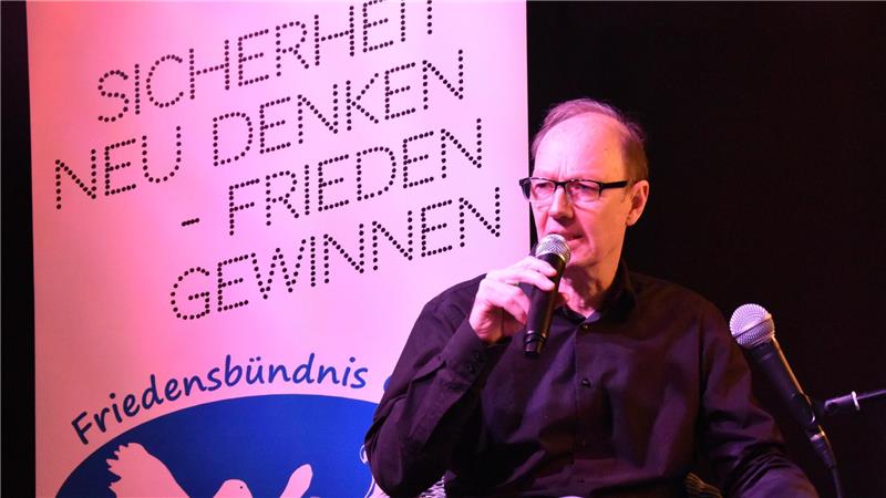 Mann mit Mikrofon vor Plakat mit den Worten 'SICHERHEIT NEU DENKEN - FRIEDEN GEWINNEN' und dem Schriftzug 'Friedensbündnis'