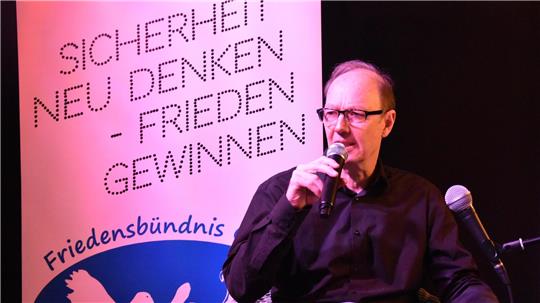 Mann mit Mikrofon vor Plakat mit den Worten 'SICHERHEIT NEU DENKEN - FRIEDEN GEWINNEN' und dem Schriftzug 'Friedensbündnis'