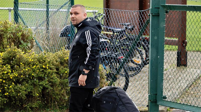Ein Mann in schwarzer Sportjacke zieht einen großen schwarzen Rollkoffer und steht vor einem grünen Maschendrahtzaun mit Fahrrädern dahinter.