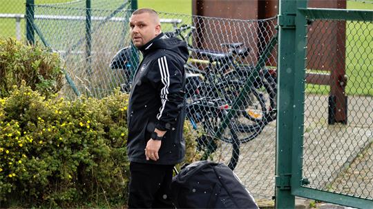 Ein Mann in schwarzer Sportjacke zieht einen großen schwarzen Rollkoffer und steht vor einem grünen Maschendrahtzaun mit Fahrrädern dahinter.