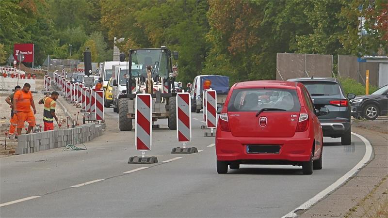 Straße mit Baustelle, mehreren Bauarbeitern in orangefarbener Kleidung, Baufahrzeugen und Autos, darunter ein rotes Fahrzeug im Vordergrund