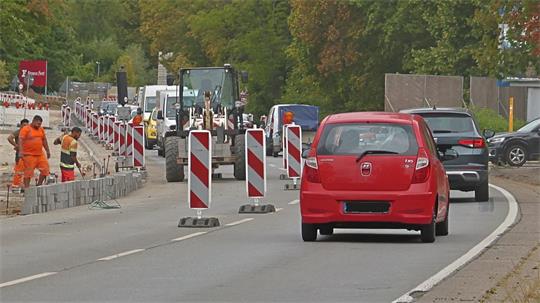 Straße mit Baustelle, mehreren Bauarbeitern in orangefarbener Kleidung, Baufahrzeugen und Autos, darunter ein rotes Fahrzeug im Vordergrund