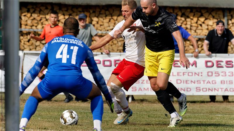 Zwei Fußballspieler in Aktion vor einem Torwart in blau auf einem Spielfeld mit Holzstapel im Hintergrund