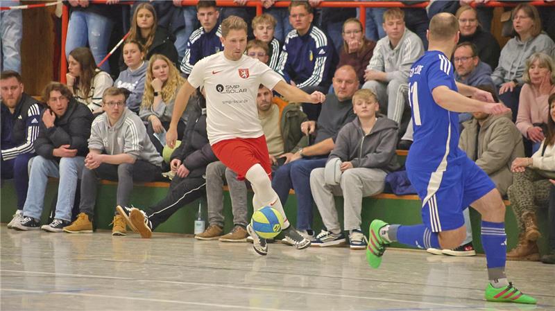 Zwei Hallenfußballspieler in Aktion, einer in weißem Trikot und roter Hose, der andere in blauem Trikot und blauer Hose, vor Zuschauern auf einer Tribüne