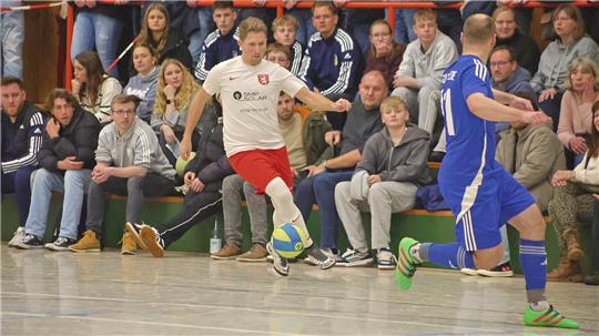 Zwei Hallenfußballspieler in Aktion, einer in weißem Trikot und roter Hose, der andere in blauem Trikot und blauer Hose, vor Zuschauern auf einer Tribüne