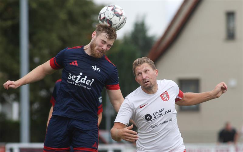 Zwei Fußballspieler springen zum Kopfball, einer trägt ein dunkelblaues Trikot mit rotem Rand, der andere ein weißes Trikot mit roter Hose