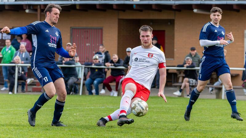 Drei Fußballspieler auf dem Spielfeld, zwei in dunkelblauen Trikots und einer in weißem Trikot mit roter Hose, der den Ball mit dem Fuß spielt