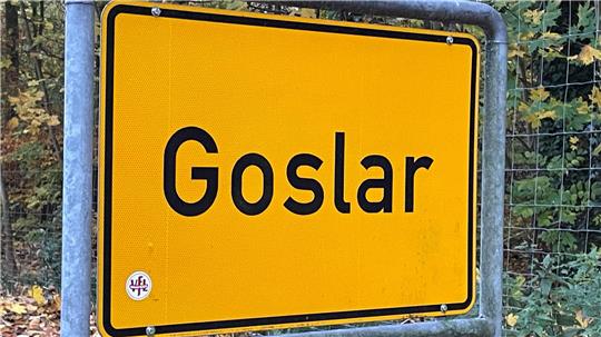 Gelbes Ortsschild mit schwarzer Aufschrift 'Goslar' an einem Metallrahmen vor herbstlichem Wald