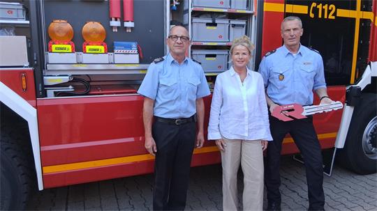 Drei Personen in Uniform und Zivilkleidung stehen vor einem roten Feuerwehrfahrzeug mit geöffneten Fächern und Ausrüstung.