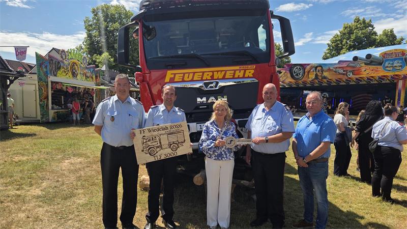 Fünf Personen stehen vor einem roten Feuerwehrfahrzeug und halten ein Schild mit der Aufschrift 'FF Wiedelah 2026 TLF 3000' und einen großen symbolischen Schlüssel.