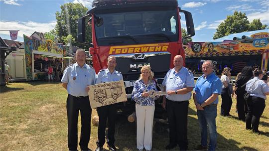 Fünf Personen stehen vor einem roten Feuerwehrfahrzeug und halten ein Schild mit der Aufschrift 'FF Wiedelah 2026 TLF 3000' und einen großen symbolischen Schlüssel.