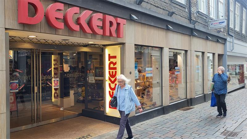 Eingang eines Geschäfts mit rotem Schriftzug "DECKERT" über der Tür und auf einem vertikalen Schild, zwei ältere Personen gehen auf dem gepflasterten Gehweg daran vorbei