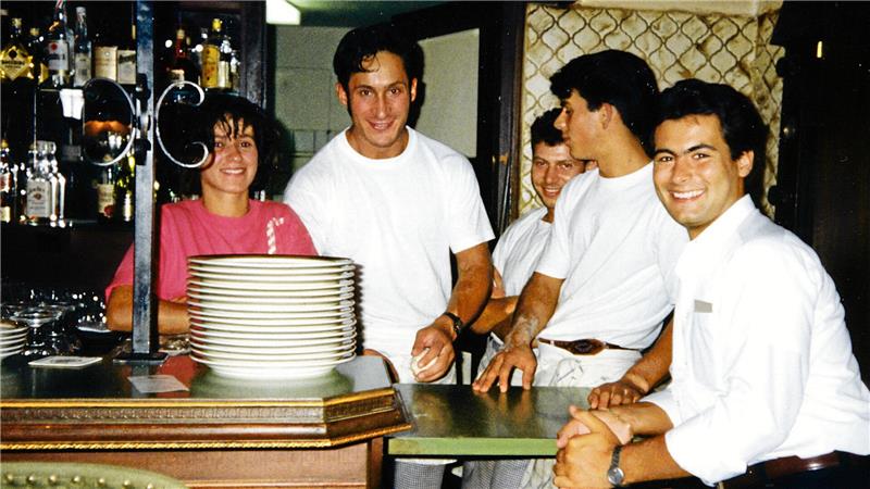 Fünf Personen stehen und sitzen an einer Bar mit gestapelten Tellern und Flaschen im Hintergrund.