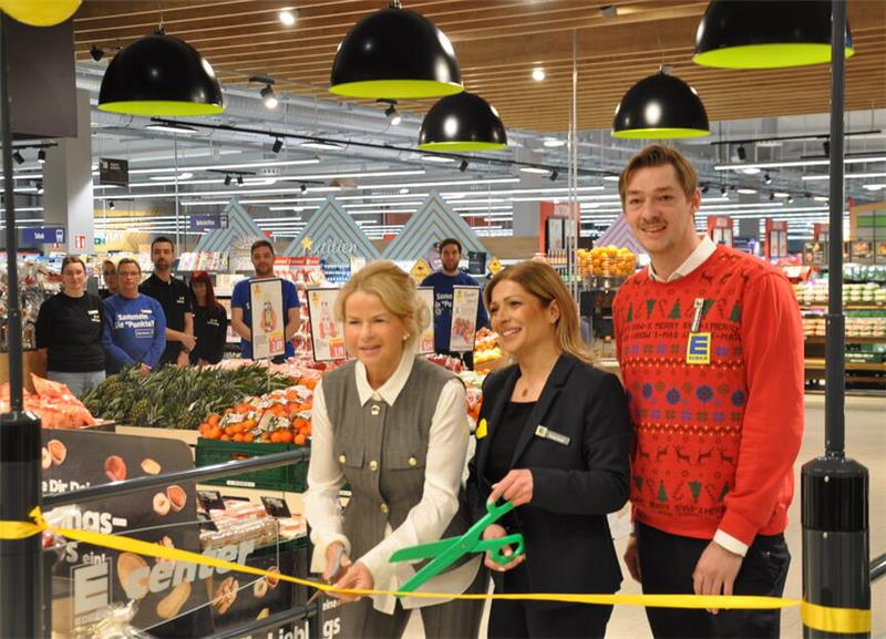 Goslarer Edeka-Center eröffnet: Erste Kunden kommen um sieben Uhr Drei Personen stehen in einem Supermarkt vor einem gelben Band, eine hält eine große grüne Schere, im Hintergrund Obst und Gemüse sowie mehrere schwarze Deckenlampen.