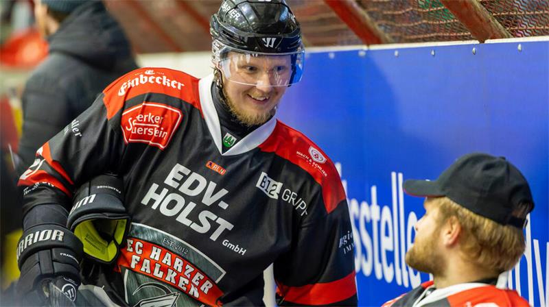 Zwei Eishockeyspieler in schwarzen und roten Trikots mit dem Schriftzug "EC Harzer Falken" unterhalten sich an der Bande.