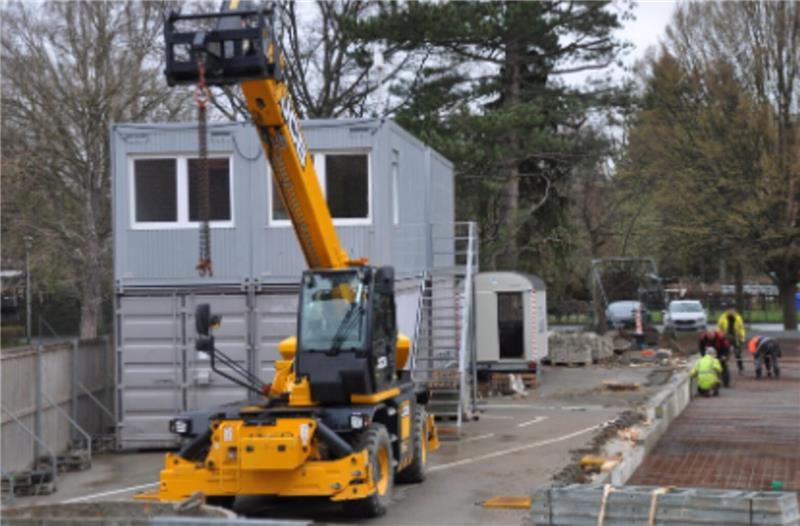 Oker drückt aufs Tempo: Millionenschwere Module rücken an Gelber Teleskoplader hebt einen grauen Container auf einer Baustelle mit mehreren Arbeitern und Fahrzeugen im Hintergrund