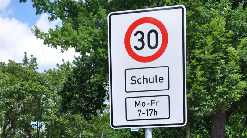 Verkehrsschild mit Geschwindigkeitsbegrenzung 30 km/h und Zusatzschild 'Schule' sowie Zeitangabe 'Mo-Fr 7-17h' vor grünem Laub