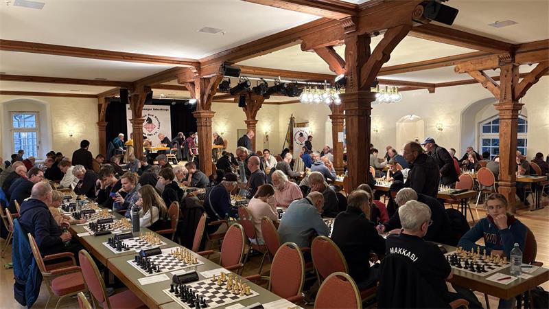 Großer Raum mit vielen Menschen, die an langen Tischen Schach spielen.
