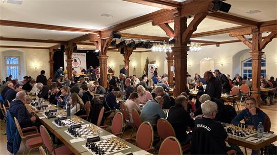 Großer Raum mit vielen Menschen, die an langen Tischen Schach spielen.