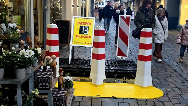 Fußgängerzone mit Absperrung aus weißen Pfosten mit roten Streifen und gelbem Boden, daneben Blumen und Deko auf dem Bürgersteig.