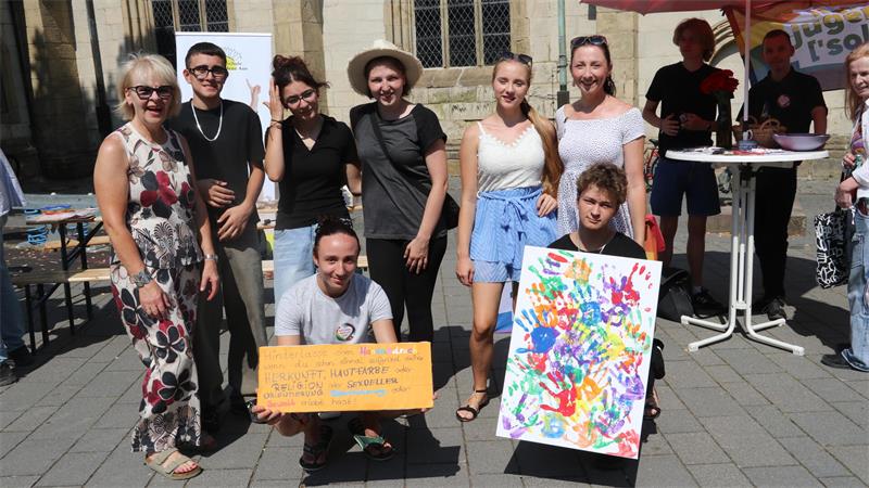 Gruppe von acht Personen vor einer Kirche, zwei halten bunte Plakate mit Handabdrücken und Text, andere stehen oder knien lächelnd zusammen