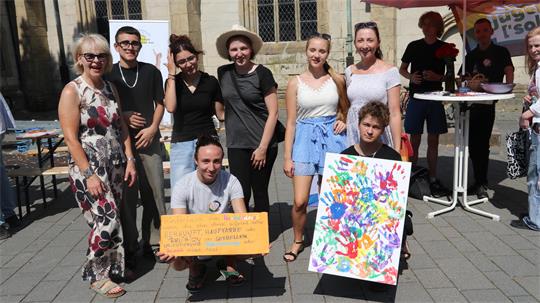 Gruppe von acht Personen vor einer Kirche, zwei halten bunte Plakate mit Handabdrücken und Text, andere stehen oder knien lächelnd zusammen