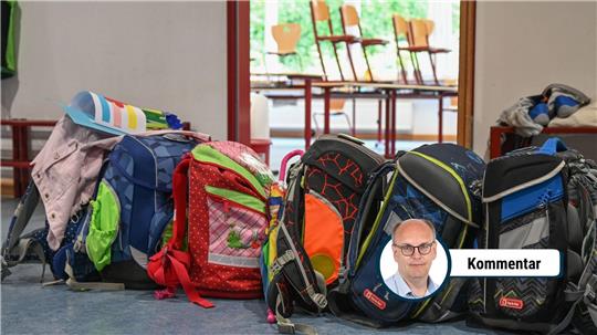 Mehrere bunte Schulranzen stehen in einer Reihe auf dem Boden eines Klassenraums, im Hintergrund sind auf Tischen gestapelte Stühle zu sehen.