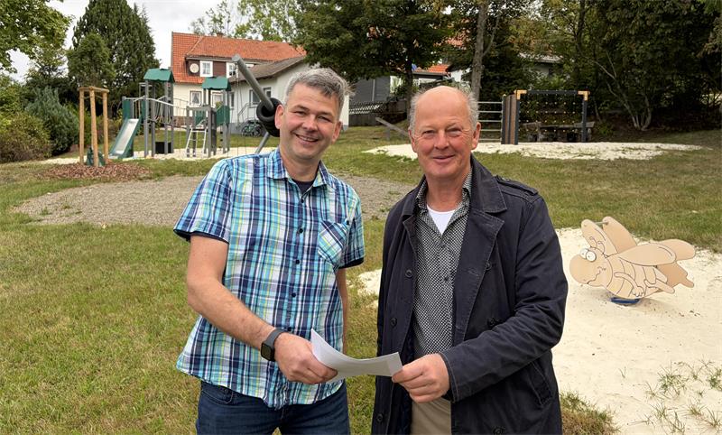 Zwei Männer stehen auf einem Spielplatz und halten einen Zettel in der Hand. 