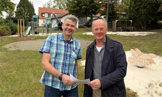 Zwei Männer stehen auf einem Spielplatz und halten einen Zettel in der Hand. 