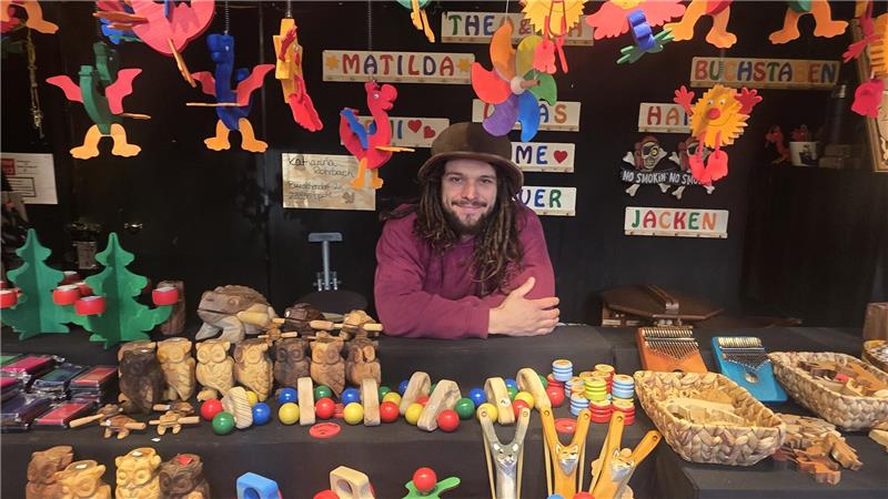 Marktstand mit handgefertigten Holzspielzeugen und Figuren, bunte hängende Holzdekorationen, Person mit Hut sitzt dahinter.