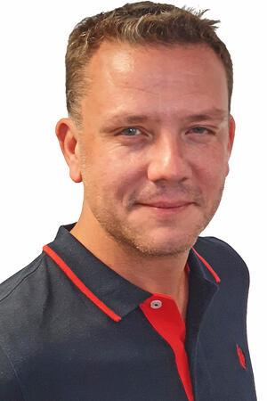 Ein Mann im blau-roten Shirt steht vor einer weißen Wand.