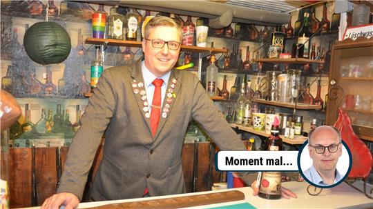 Mann in grauem Anzug mit roter Krawatte und Anstecknadeln steht hinter einer Bar mit verschiedenen Flaschen und Gläsern im Hintergrund