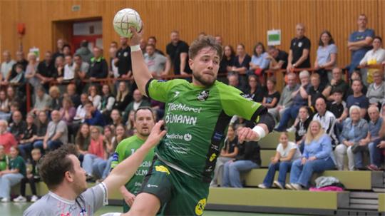 Handballspieler in grüner Sportkleidung springt mit Ball in der Hand über einen Gegner in grauer Sportkleidung in einer Halle mit Zuschauern auf der Tribüne