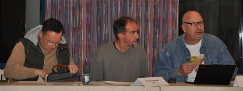 Drei Männer sitzen an einem Tisch mit Mikrofon, Namensschild und Laptop vor einem Vorhang