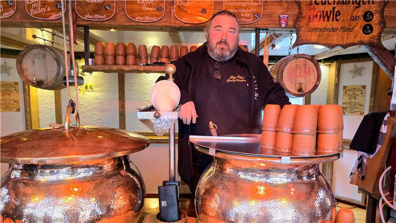 Verkäufer steht hinter zwei großen kupfernen Kesseln mit Holzgriffen, daneben Stapel brauner Tonbecher und hängende Schilder mit Getränkenamen.