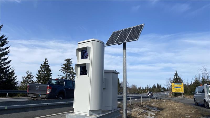 Geschwindigkeitsmessgerät mit Solarpanel an einer Straße mit Fahrzeugen und Bäumen im Hintergrund.