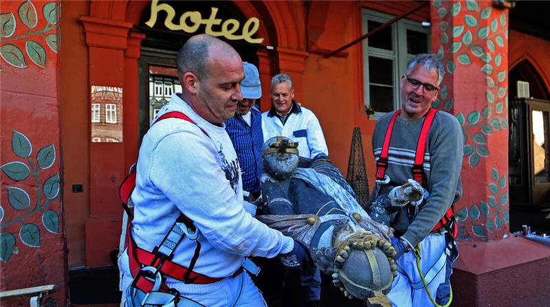 Vier Personen mit Sicherheitsgurten heben gemeinsam eine große Statue vor einem rot gestrichenen Gebäude mit dem Schriftzug 'hotel'