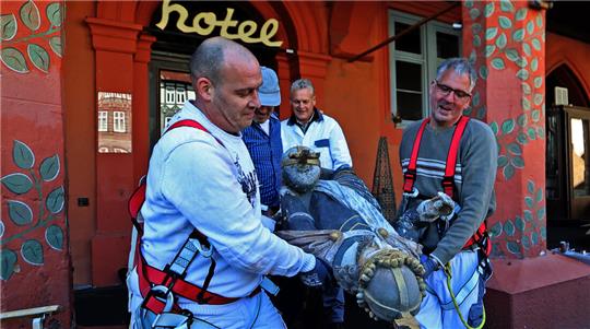 Vier Personen mit Sicherheitsgurten heben gemeinsam eine große Statue vor einem rot gestrichenen Gebäude mit dem Schriftzug 'hotel'