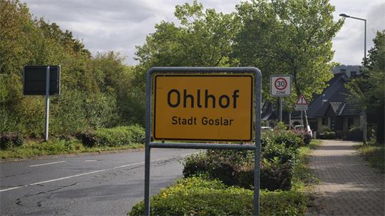 Ortseingangsschild von Ohlhof, einem Stadtteil von Goslar. Im Hintergrund sind eine Straße, Bäume, Wohnhäuser sowie Verkehrszeichen für eine Tempo-30-Zone und eine geänderte Vorfahrt zu sehen.