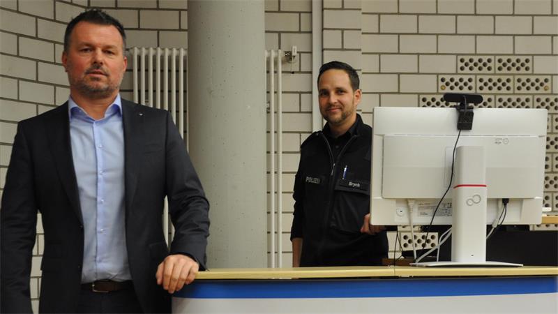 Zwei Männer stehen hinter einem Tresen, einer trägt Anzug, der andere eine Uniform, im Hintergrund ein weißer Heizkörper und ein Computerbildschirm mit Webcam