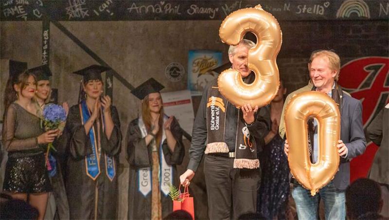 Gruppe von Menschen in festlicher Kleidung auf einer Bühne, zwei halten große goldene Luftballons in Form der Zahlen 3 und 0