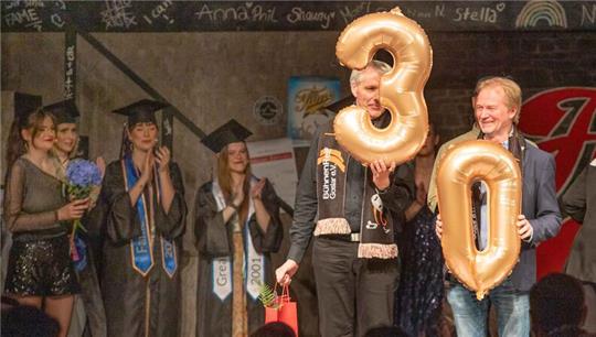 Gruppe von Menschen in festlicher Kleidung auf einer Bühne, zwei halten große goldene Luftballons in Form der Zahlen 3 und 0