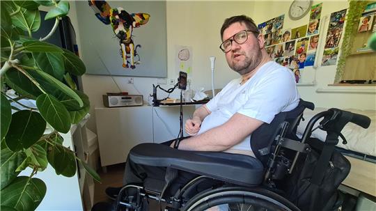 Ein Mann sitzt in einem Zimmer im Rollstuhl. Um ihn herum Bilder, Fotos und eine Grünpflanze.
