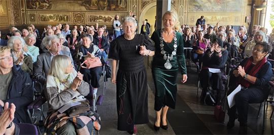 Zwei Frauen haben sich untergehakt und schreiten durch eine Menge sitzender Zuschauer in einem herrschaftlichen Raum. 