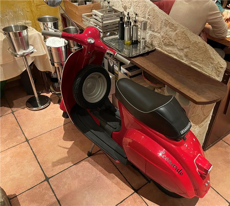 Roter Motorroller mit schwarzem Sitz steht in einem Raum mit Fliesenboden neben einem Holztresen und einem Tisch mit Stühlen.
