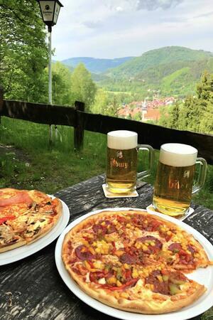 Zwei Biergläser und zwei Pizzen auf einem Holztisch mit Blick auf bewaldete Hügel und ein Dorf im Tal.