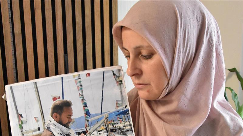 Frau mit hellrosa Kopftuch hält ein Schild mit einem Foto eines Mannes vor einem Hafen mit Segelbooten